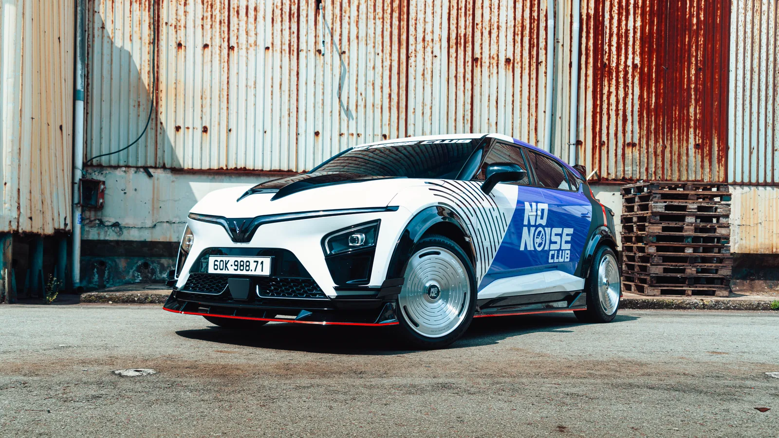 The real VinFast VF7 with the No Noise Club SEED body kit and livery, parked by a rusted warehouse wall